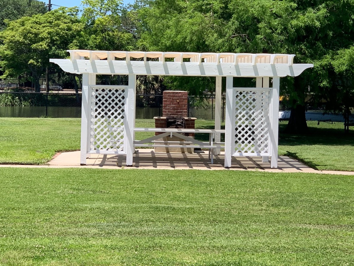 A white pergola with lattice sides stands in a sunny park, surrounded by lush green grass.