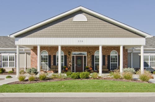 A suburban residential building with brick facade and white columns, surrounded by a landscaped front yard.