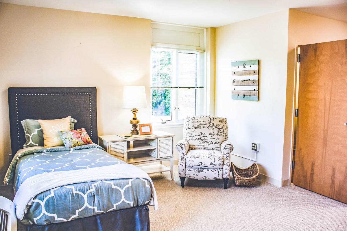 A cozy bedroom with a neatly made bed, an armchair, and a lamp by the window.