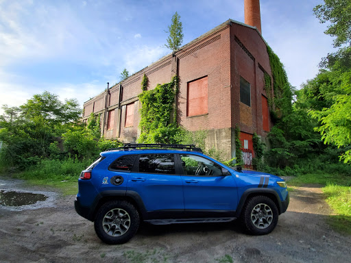 A blue SUV parked next to an overgrown, abandoned brick building under a clear sky.