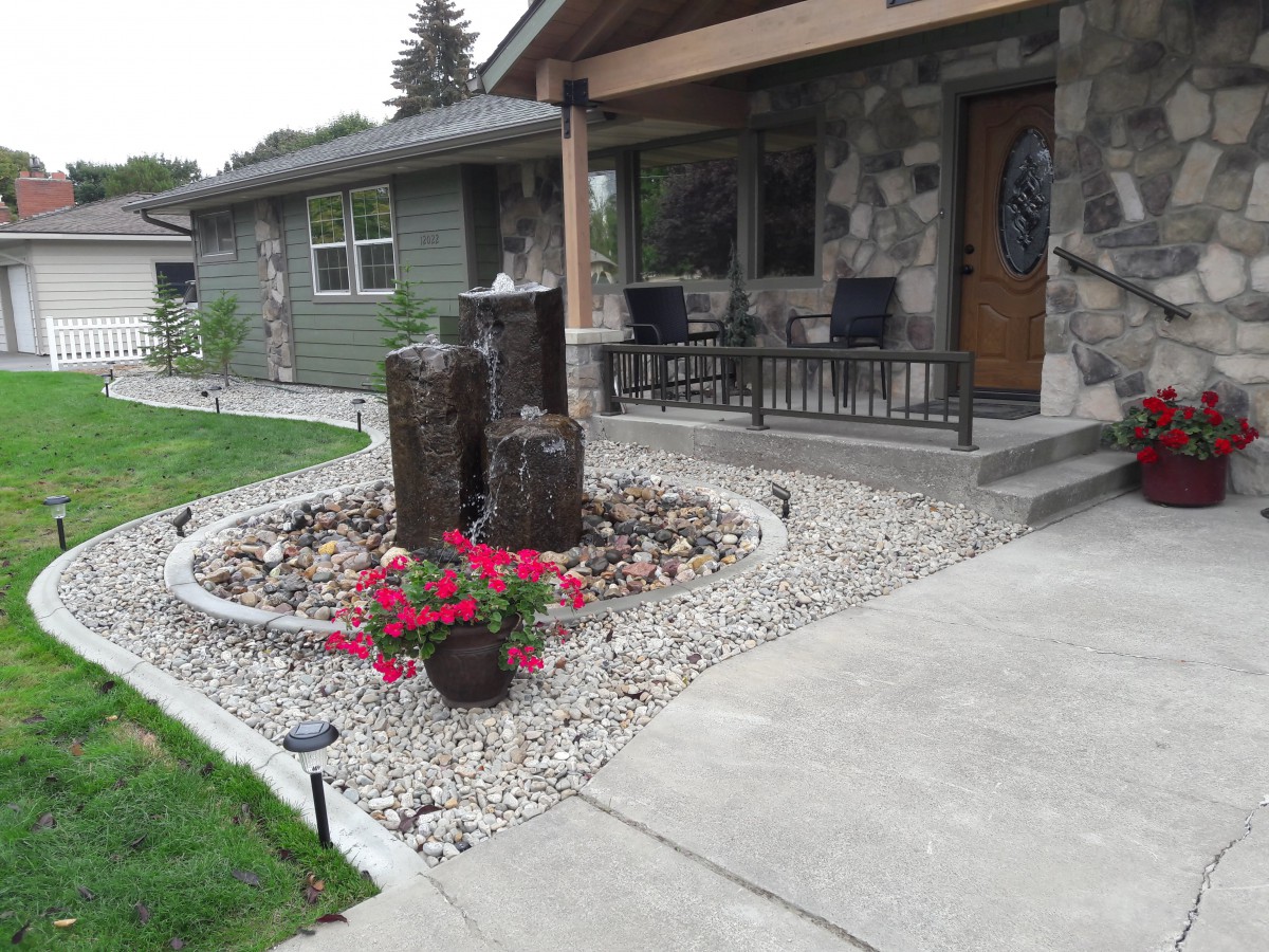 A well-maintained front yard featuring a rock garden with a fountain and vibrant flowers, adjacent to a stone house with outdoor seating.