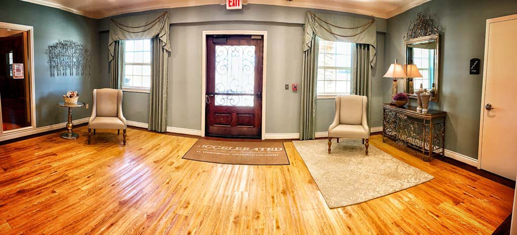 A cozy room with polished wooden floors, a central door flanked by windows with drapes, and an armchair on a rug.