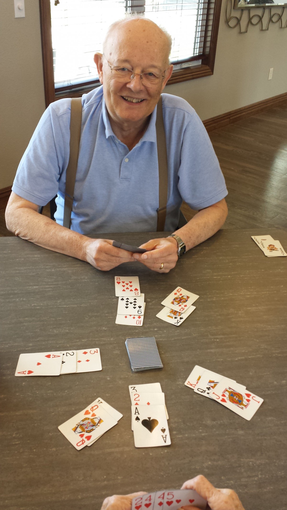 An elderly person sitting at a table playing cards, with a partially revealed hand and some cards laid out on the table.