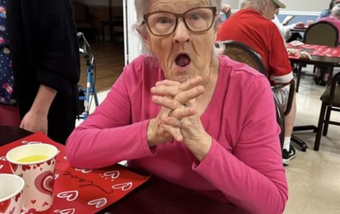 An elderly woman in glasses and a pink shirt with a surprised expression.