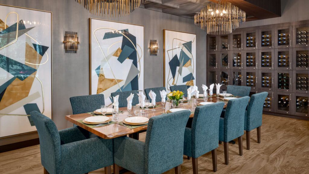 A modern dining room setting with a large table, blue chairs, abstract artwork, and elegant chandeliers.