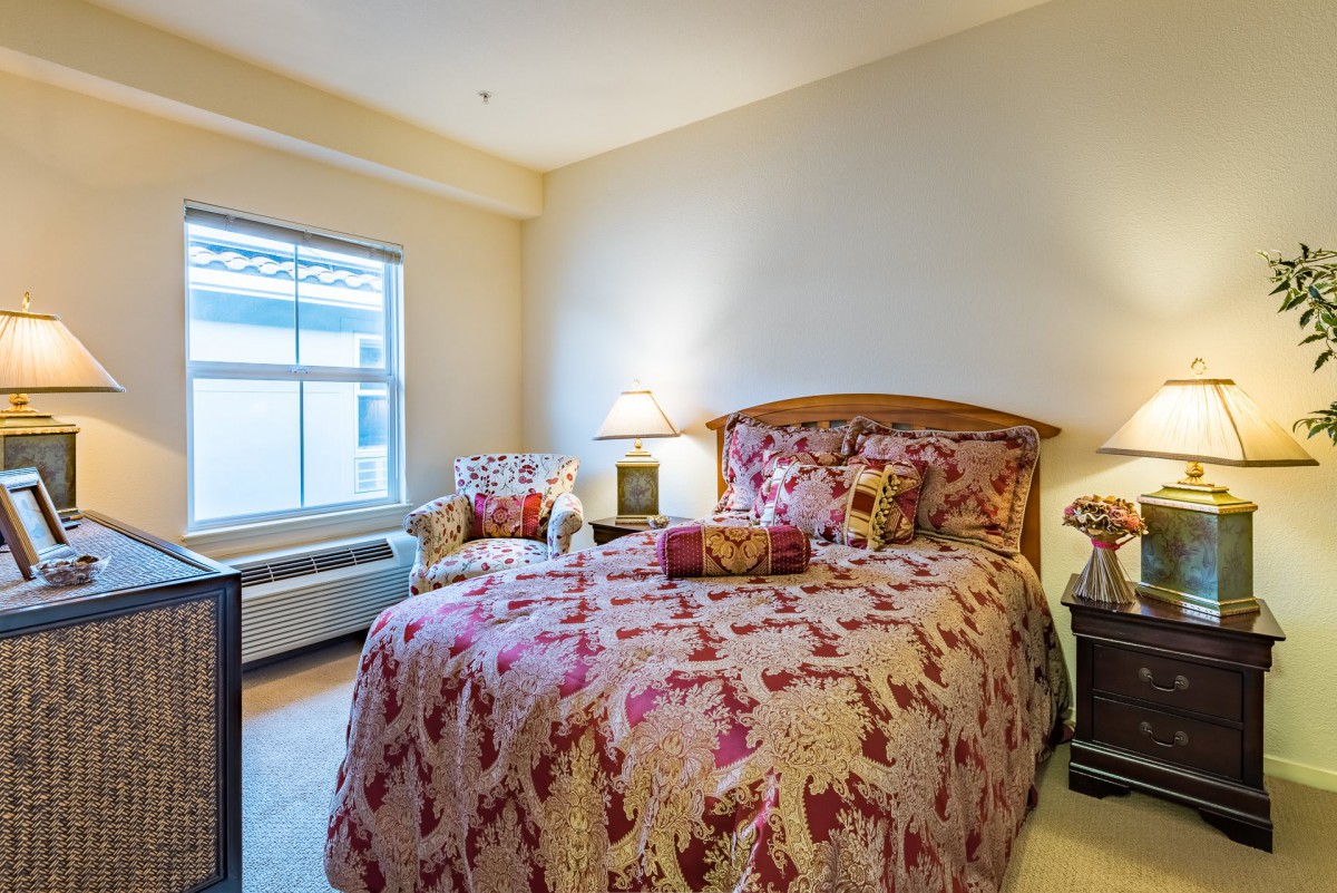 A cozy bedroom featuring a bed with floral bedding, a side table with lamps, and a window allowing natural light to brighten the space.
