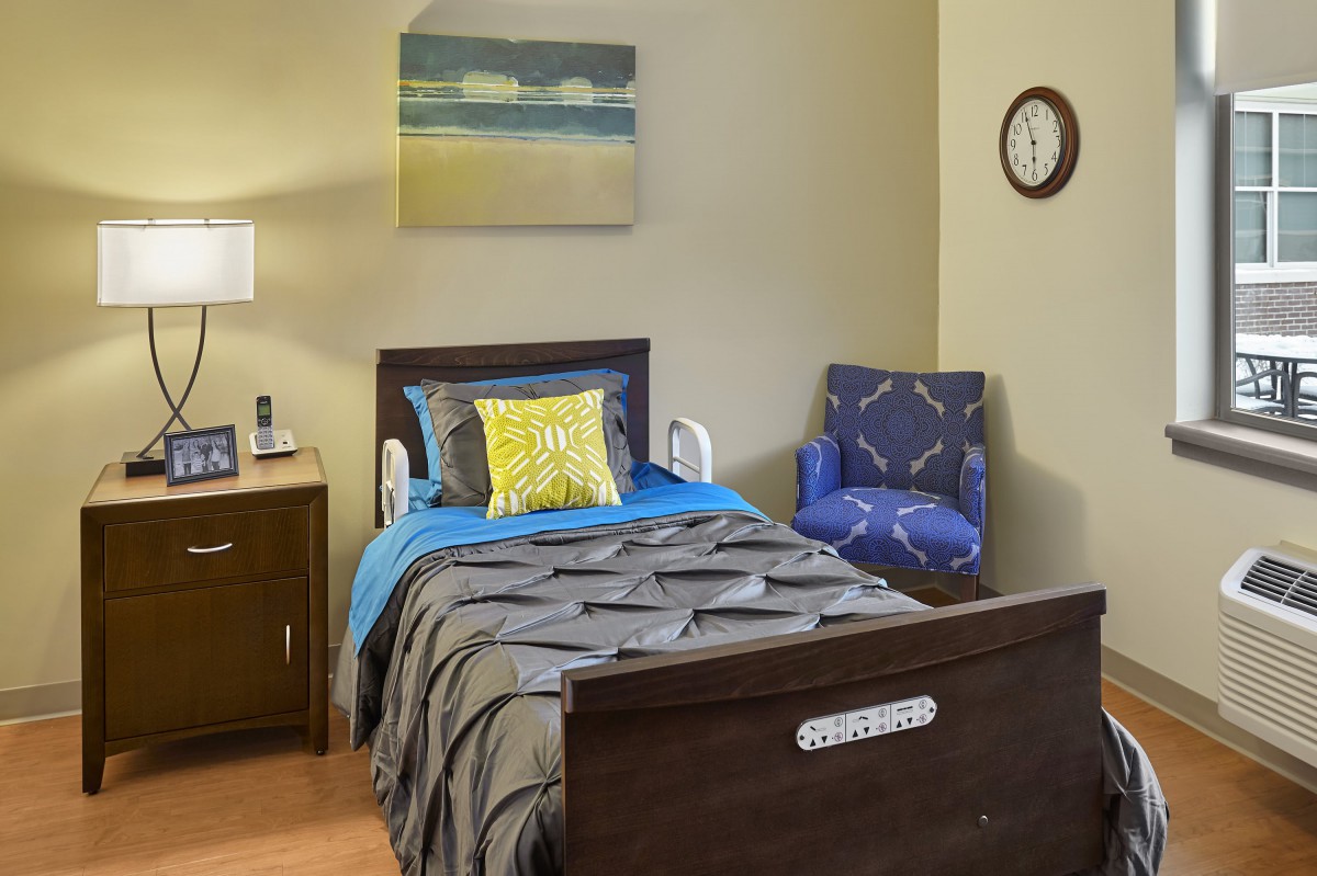 A cozy bedroom featuring a single bed with colorful pillows, a side table with a lamp, and a comfy chair next to it. A wall clock and painting decorate the space.