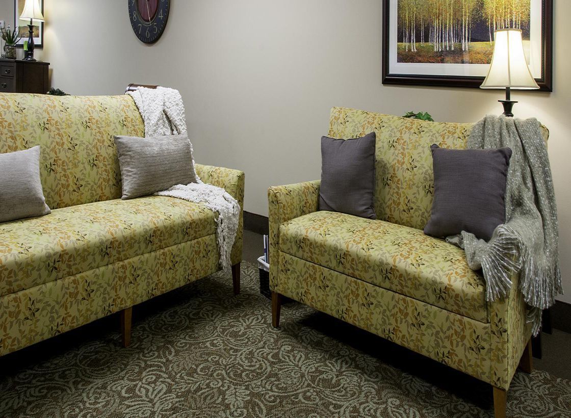 A cozy living room with two patterned sofas, accented by grey cushions and a soft throw blanket. The space is complemented by a carpet, a lamp for lighting, and wall art.
