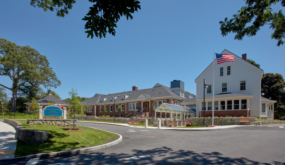 A hospital building with a flag at the entrance and a parking area outside.