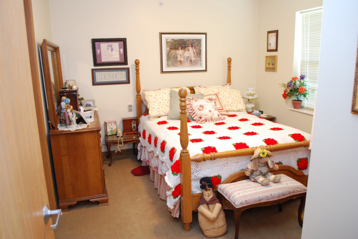 A cozy bedroom with a wooden bed featuring a red and white quilt, surrounded by framed artwork and decorative items.