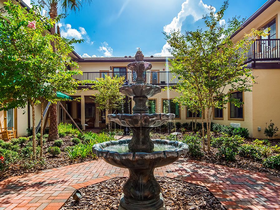 A tranquil courtyard featuring a central fountain surrounded by lush greenery and a two-story building under a bright blue sky.