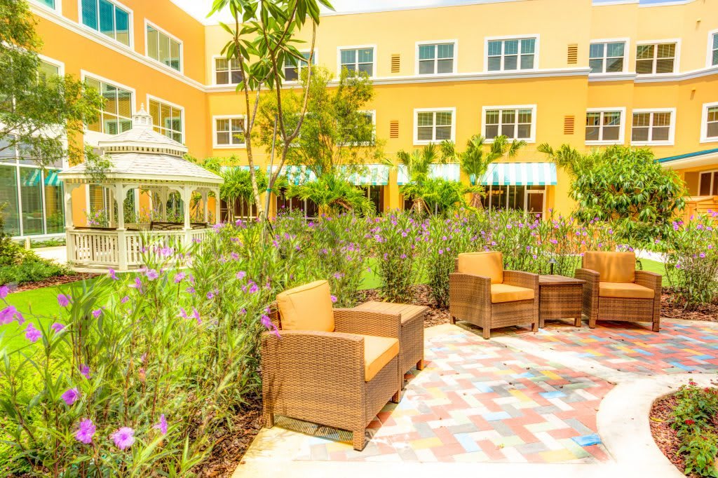 A vibrant patio area featuring wicker chairs surrounded by lush greenery and a yellow building backdrop.