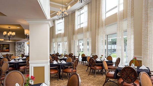 An elegant dining area with large windows and chandeliers, featuring neatly arranged empty tables.