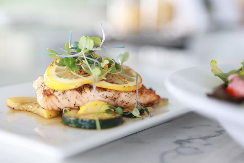 A beautifully plated dish featuring salmon topped with lemon slices and microgreens, accompanied by zucchini.