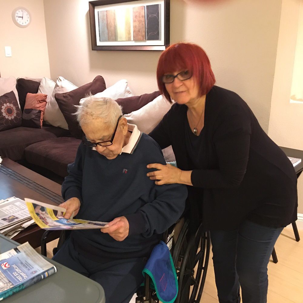 A woman with red hair stands next to an elderly person in a wheelchair who is reading a magazine in a cozy living room.