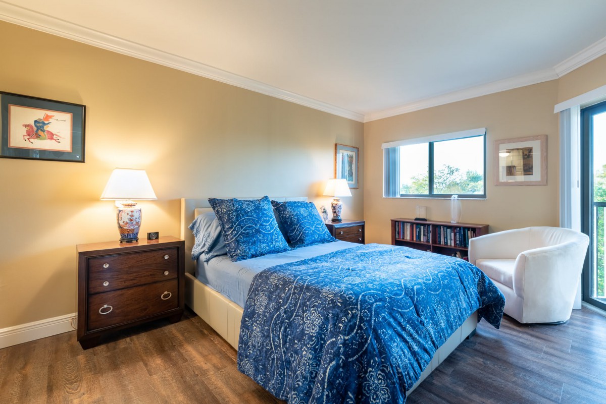 A cozy bedroom featuring a bed with blue bedding, accompanied by nightstands and lamps on either side. A window allows natural light to illuminate the space, while a chair and bookshelf add to the room's decor.