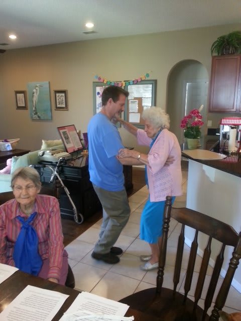 An elderly woman and a younger man dance together in a cozy living room while another woman sits nearby, smiling.