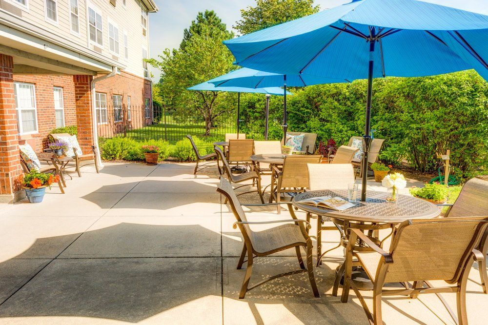 A sunny patio with tables, chairs, and blue umbrellas surrounded by greenery.