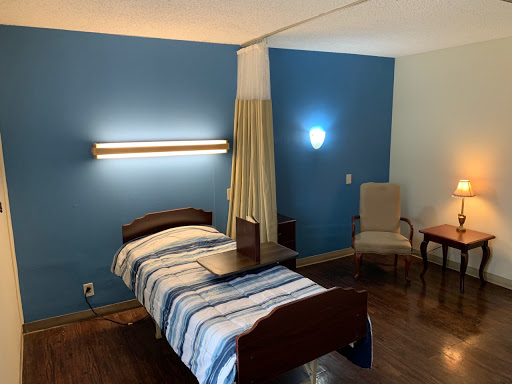 A well-lit hospital room featuring a single bed with striped bedding, accompanied by a chair and side table.