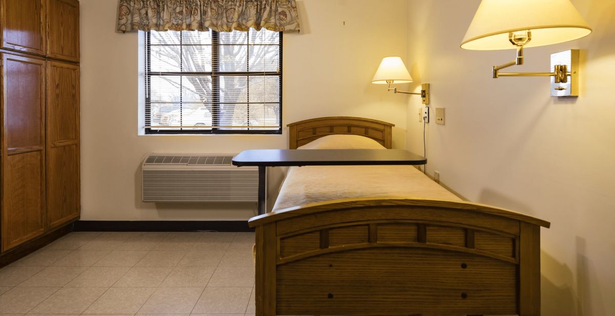 A hospital room with a single bed and a bedside lamp near a window.