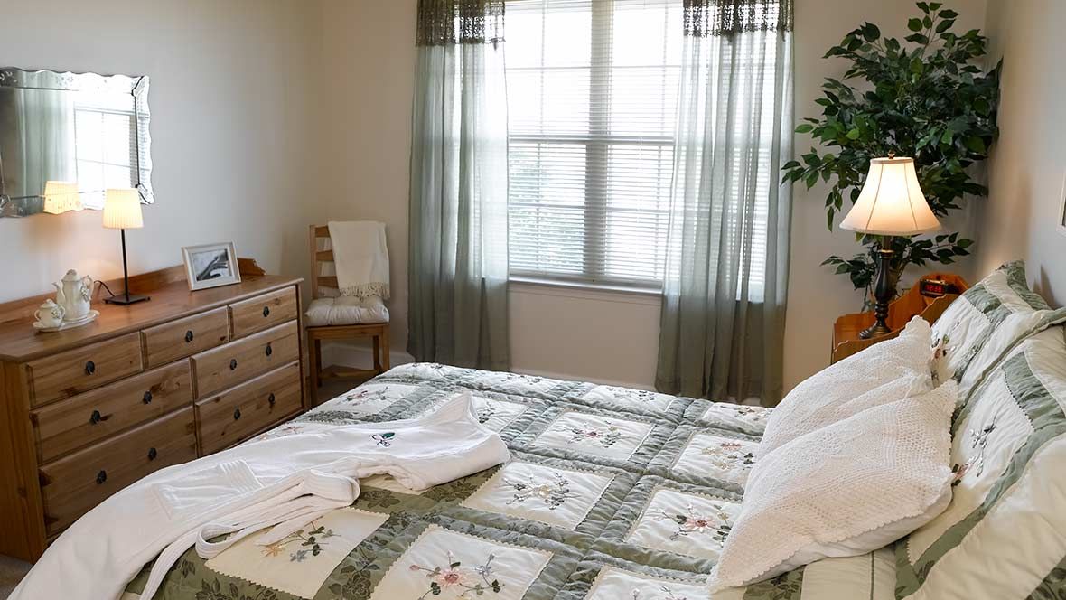 A cozy bedroom featuring a neatly made bed with floral-patterned bedding, a wooden dresser, and a chair near the window with sheer curtains.