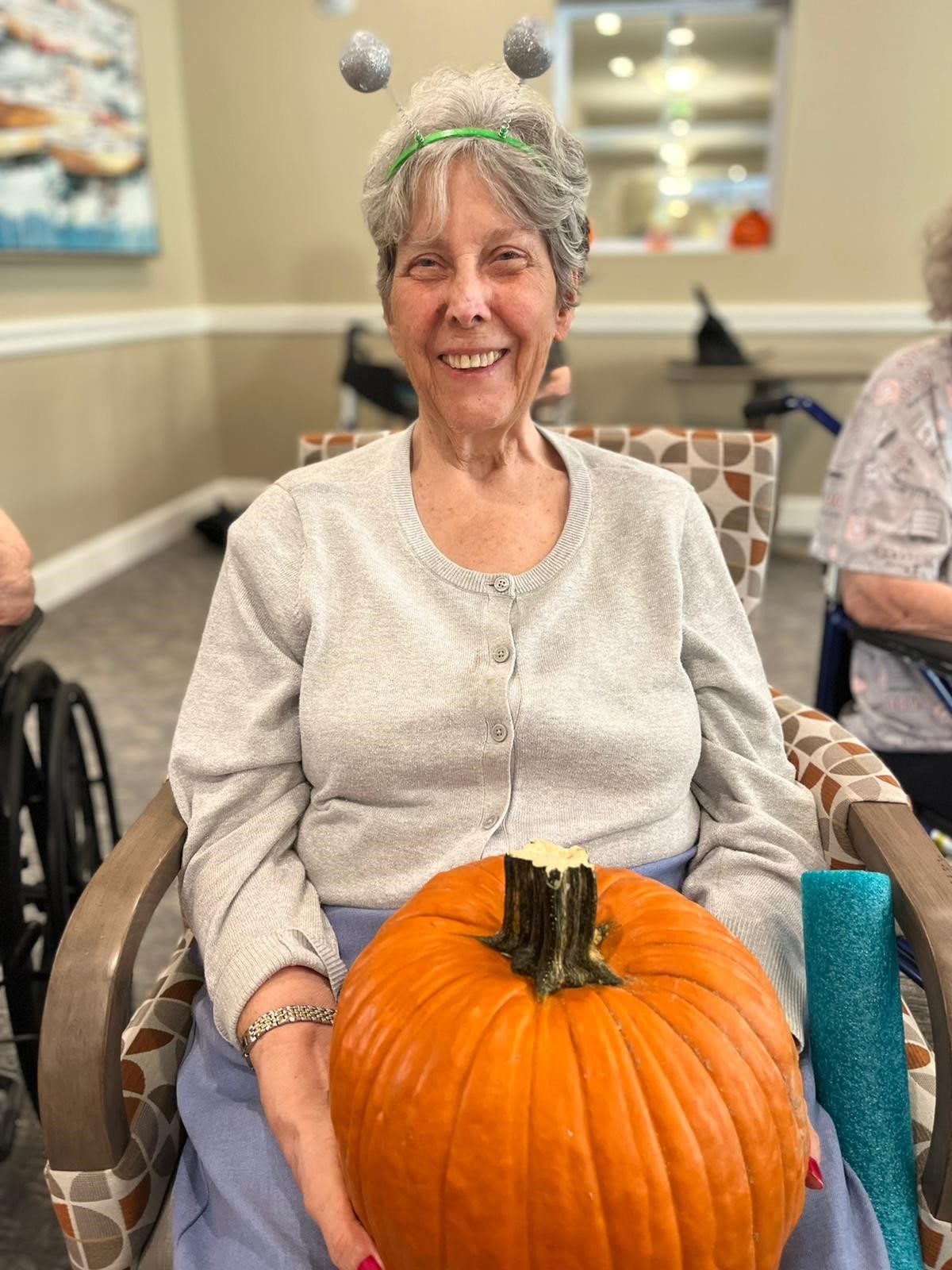 An elderly woman smiling while holding a pumpkin indoors.