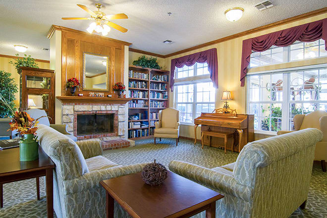A cozy living room with a fireplace and bookshelves, featuring armchairs and a piano by the window.