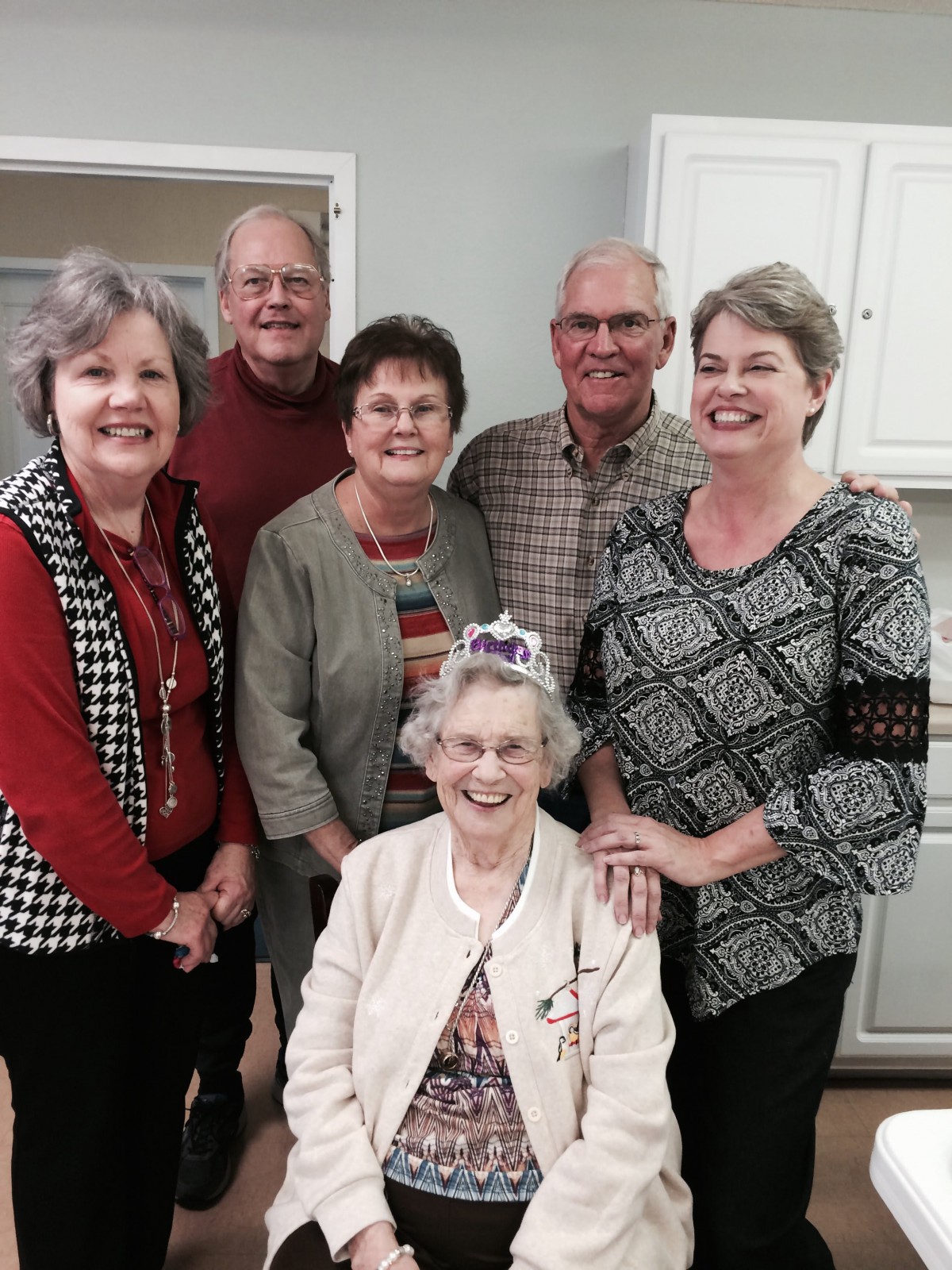 A group of smiling adults gathers around an elderly woman wearing a birthday crown.