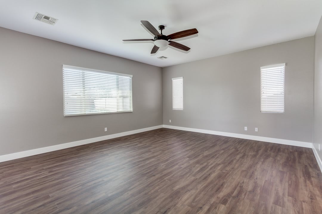 A spacious empty room with wood flooring, a ceiling fan, and ample natural light from multiple windows.