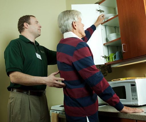 A caregiver assists an elderly man in reaching for an item in a kitchen cabinet.