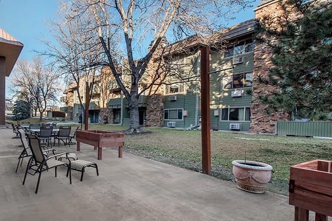 A view of an apartment building with a spacious patio area featuring chairs and trees.