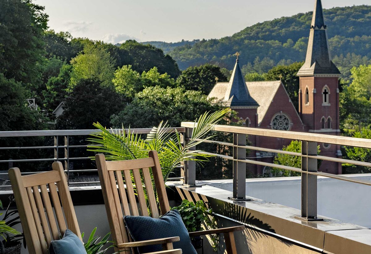 A serene balcony with wooden chairs overlooking a scenic view of trees, hills, and a church in the distance.
