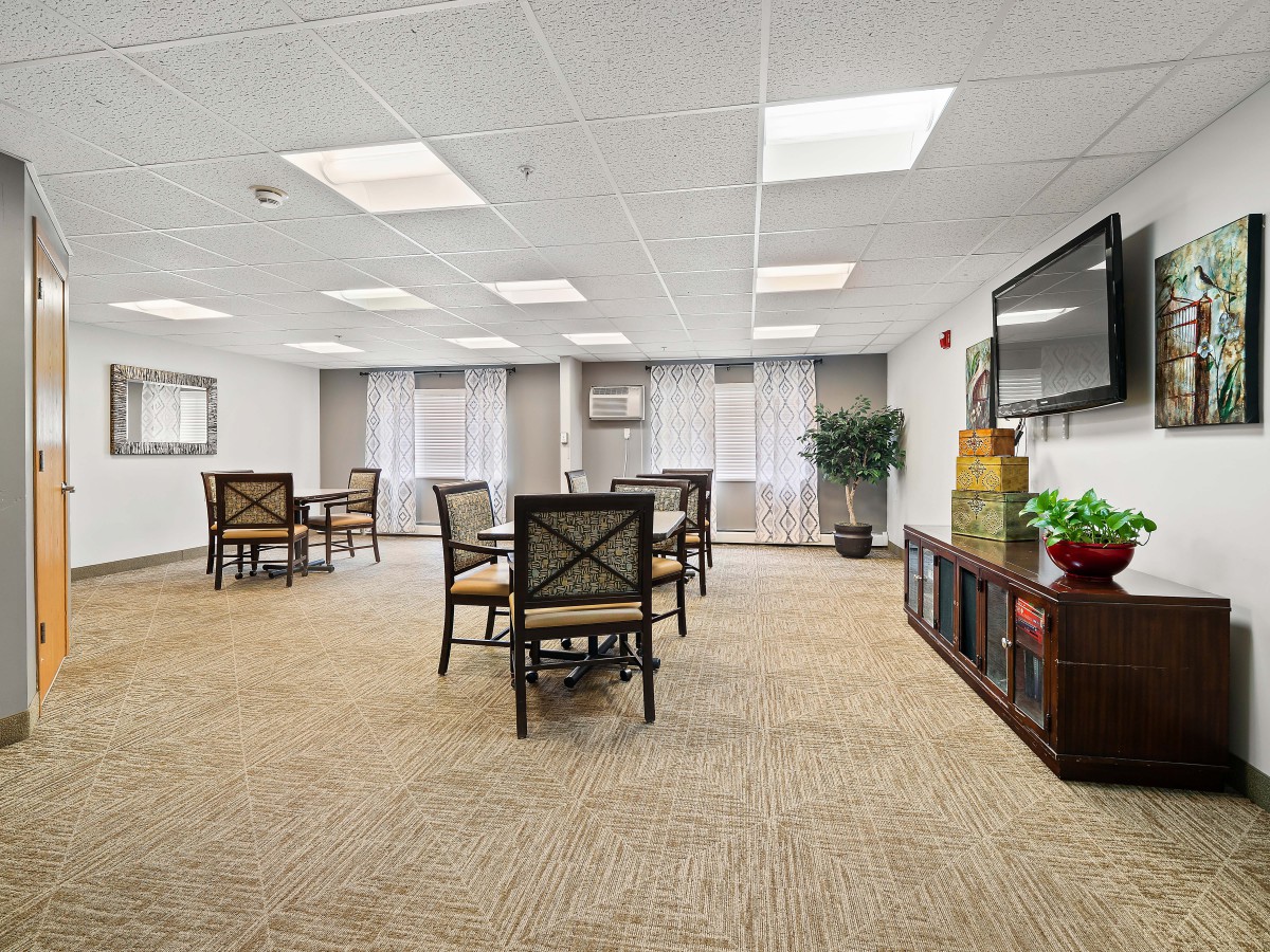 A spacious and well-lit room with tables and chairs, featuring a carpeted floor and large windows.
