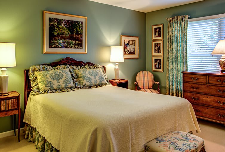 A cozy bedroom with classic decor, featuring a comfortable bed, side table with lamp, paintings on the wall, and floral curtains.