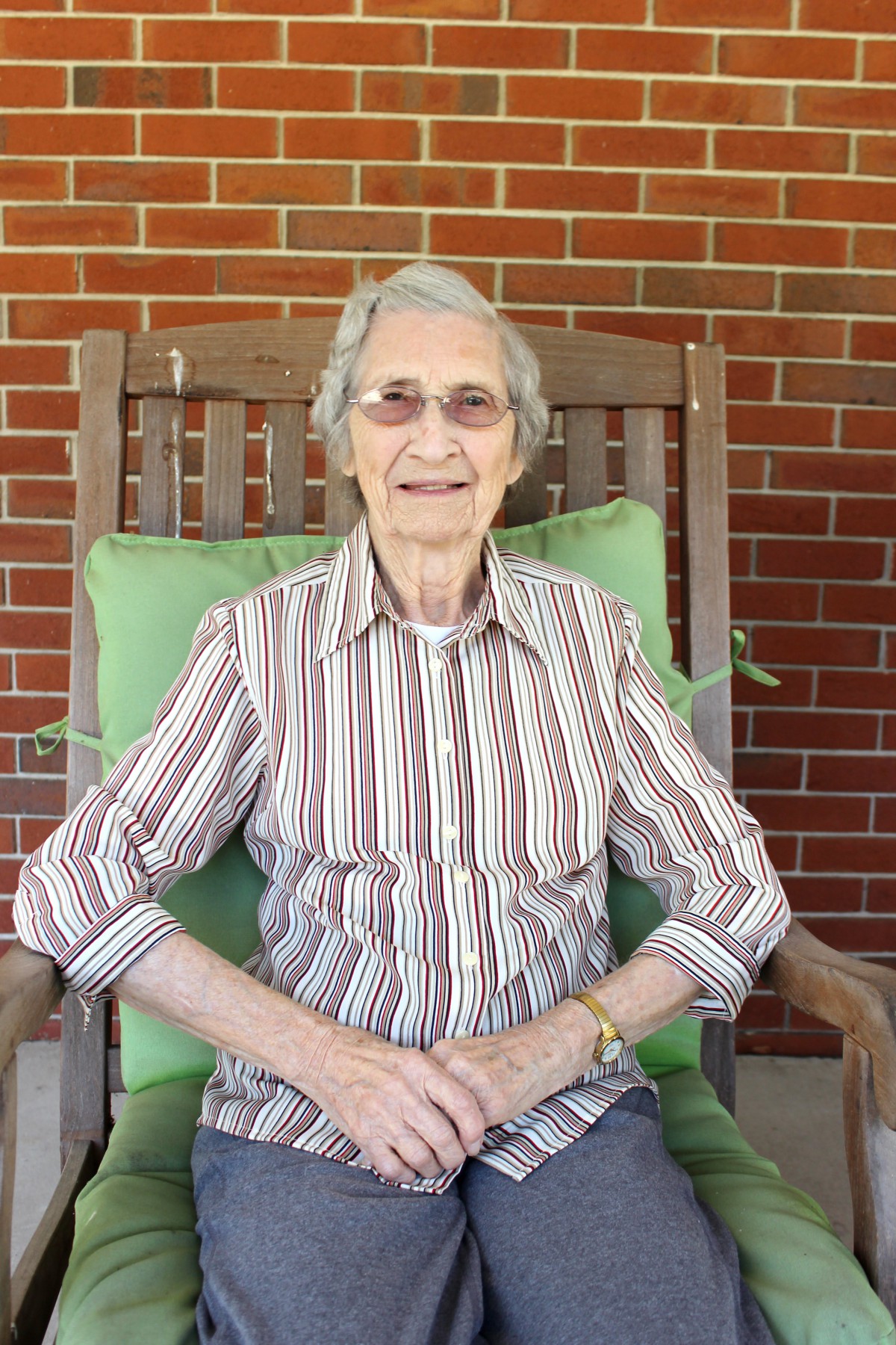 An elderly woman sitting in a wooden rocking chair against a brick wall, wearing a striped shirt and glasses.