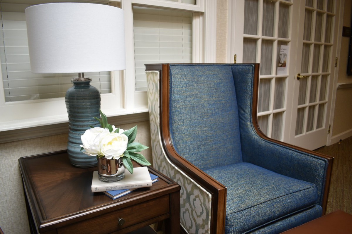 A cozy interior setting with an upholstered chair, a side table with a lamp, and decorative flowers.