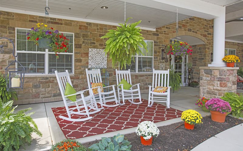 A cozy porch with white rocking chairs, a decorative rug, and vibrant plants creating a welcoming outdoor space.