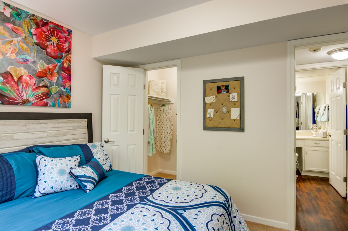 A cozy bedroom featuring blue patterned bedding, vibrant artwork on the wall, and a glimpse into an adjacent closet through an open door.