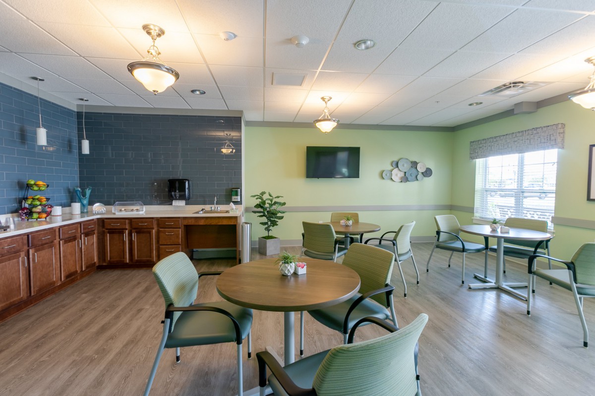 A modern dining area with a kitchenette, featuring round tables, cushioned chairs, and a wall-mounted television.
