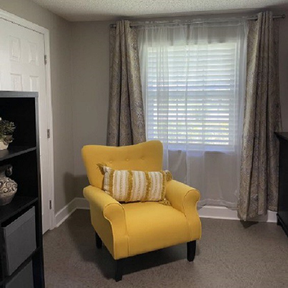 A cozy living room corner featuring a bright yellow chair, positioned beside a window with light curtains. A black shelf stands nearby, adding a touch of decor to the space.