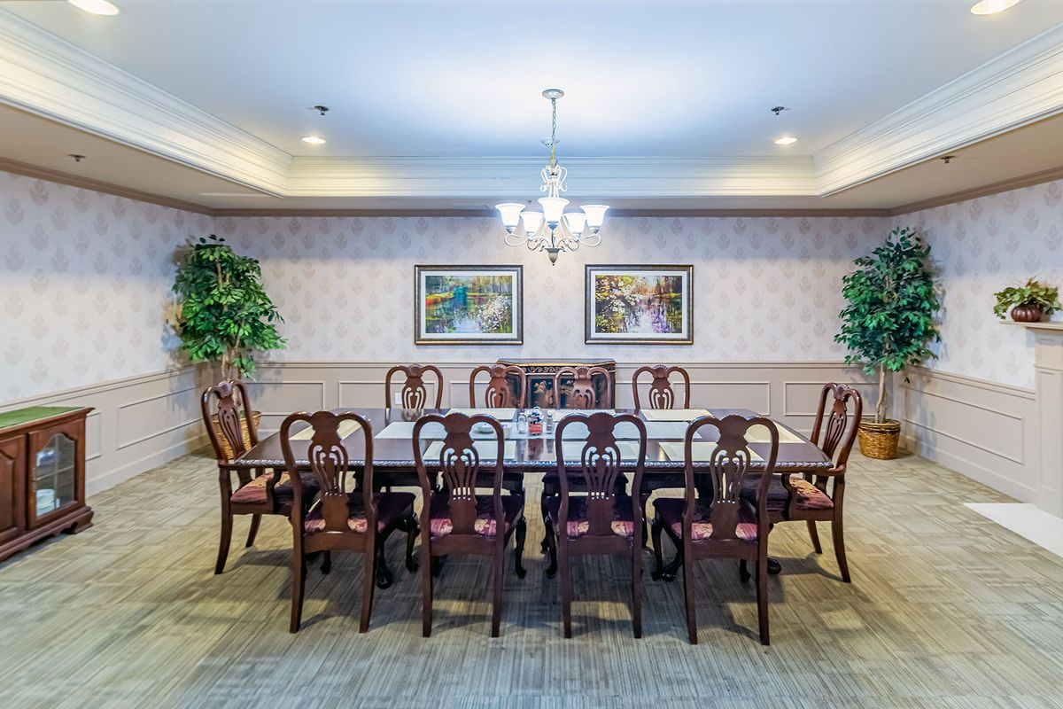A formal dining room with a long table, surrounded by chairs and illuminated by a chandelier, complemented by decorative paintings and plants.