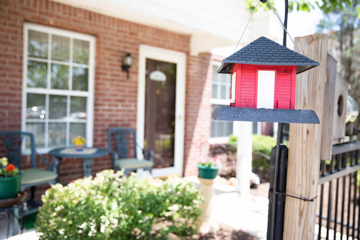 A suburban porch with a red birdhouse and outdoor seating, surrounded by greenery.