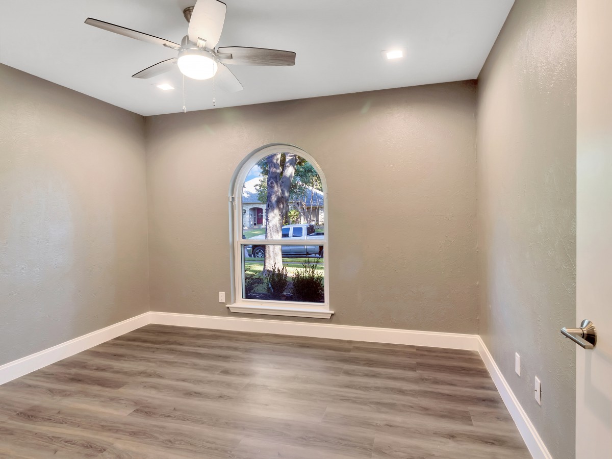 A modern empty room featuring a ceiling fan, wooden floor, and an arched window with a view outside.