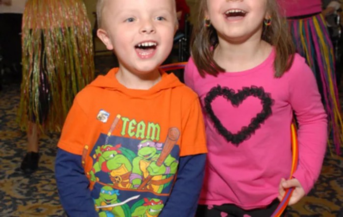 A boy and a girl smiling happily indoors, wearing colorful clothing.