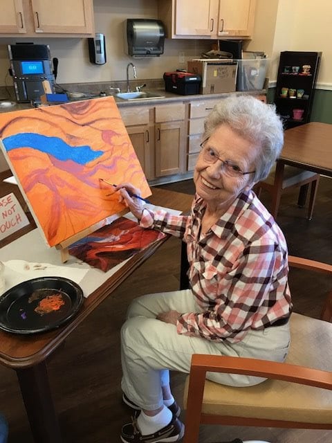 An elderly woman painting a colorful abstract artwork indoors.