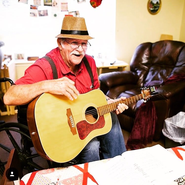 A man wearing a hat plays an acoustic guitar in a cozy living room.