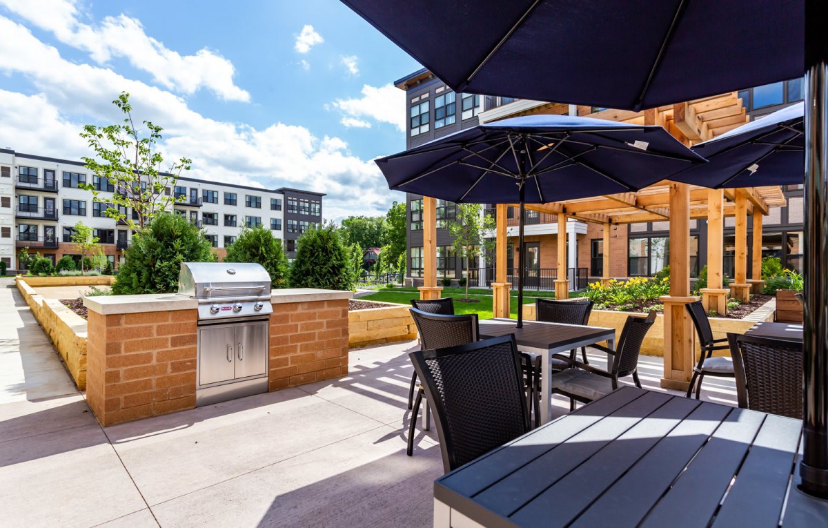 A sunny outdoor patio area with seating, umbrellas, and a BBQ grill in an apartment complex.