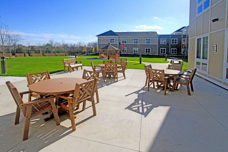 A sunlit outdoor patio area with wooden tables and chairs, adjacent to a well-maintained lawn and building.