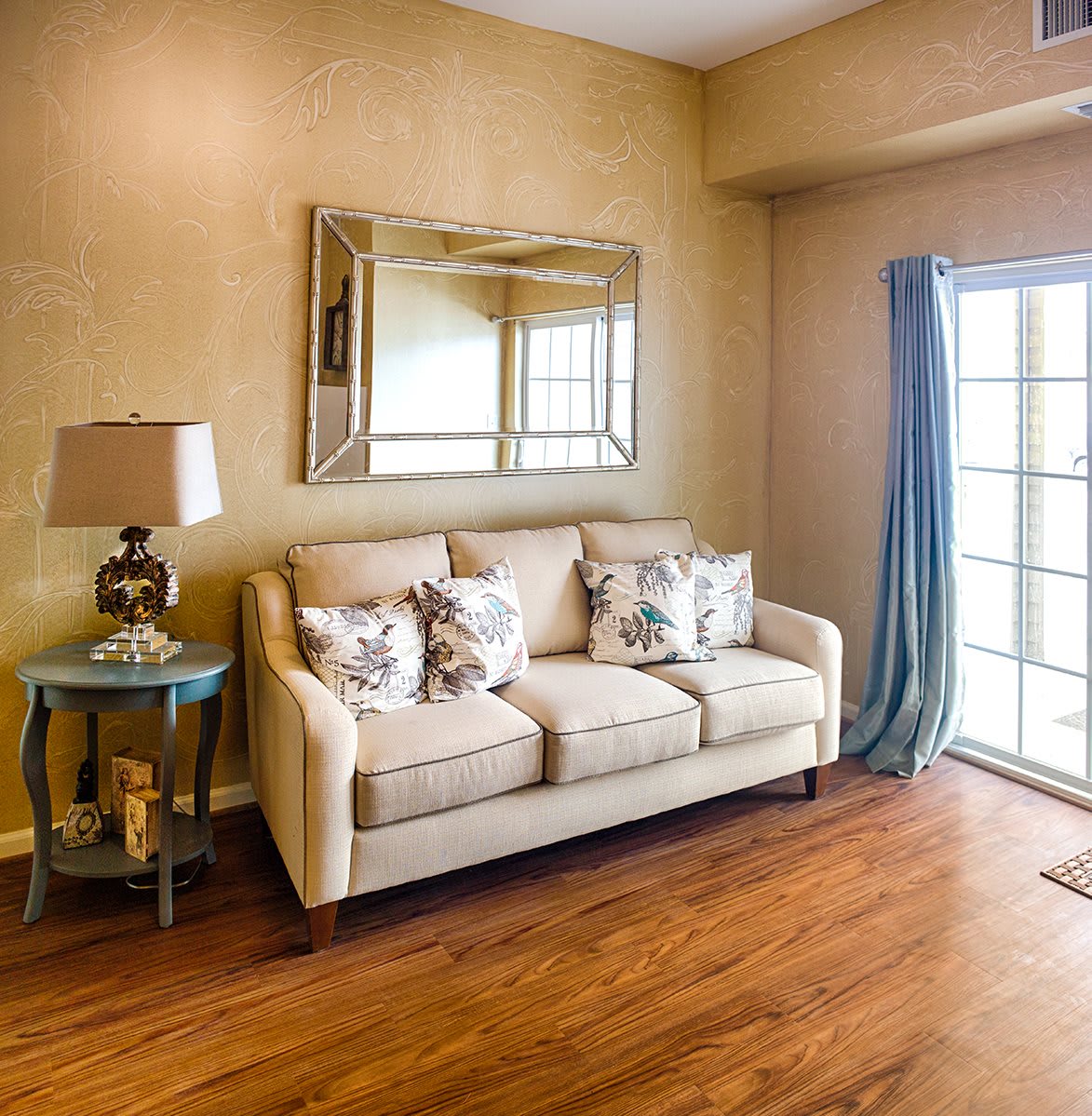 A cozy living room with a beige sofa, decorative pillows, and a side table with a lamp, featuring wooden flooring and a large window.