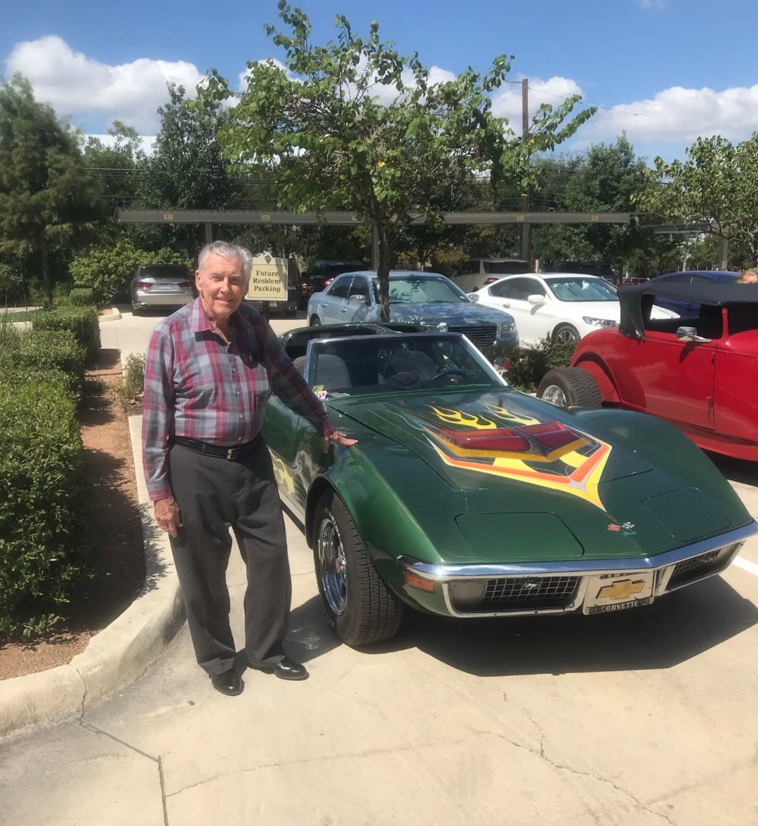 A man stands next to a vintage car in a parking lot on a sunny day.